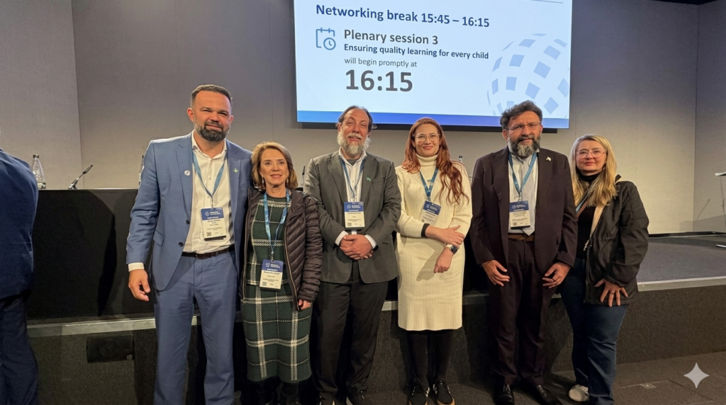 Comitiva de educadores e autoridades brasileiras posa para foto no Fórum Mundial de Educação em Londres. O grupo de seis pessoas veste trajes formais e crachás oficiais do evento, posando alinhado à frente de um grande painel digital de fundo azul que exibe informações sobre a plenária "Ensuring quality learning for every child".
