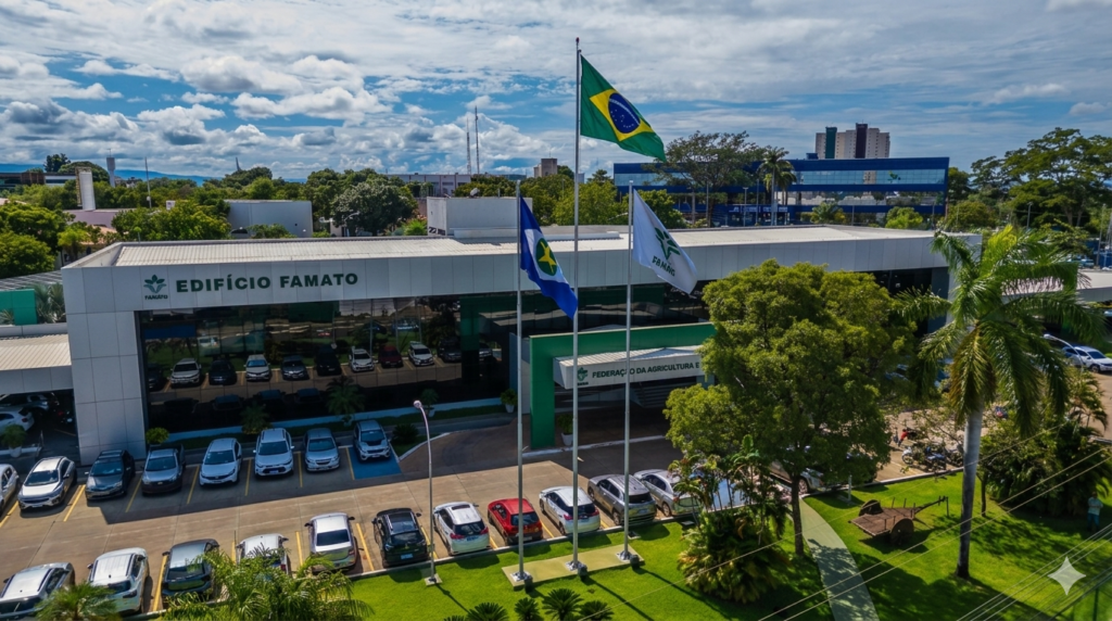 Vista aérea da fachada do Edifício Famato em Cuiabá, com as bandeiras do Brasil, de Mato Grosso e da Famato hasteadas. A imagem destaca a sede da Federação da Agricultura e Pecuária de Mato Grosso sob céu com nuvens, ideal para ilustrar notícias sobre o agronegócio e legislação tributária estadual.