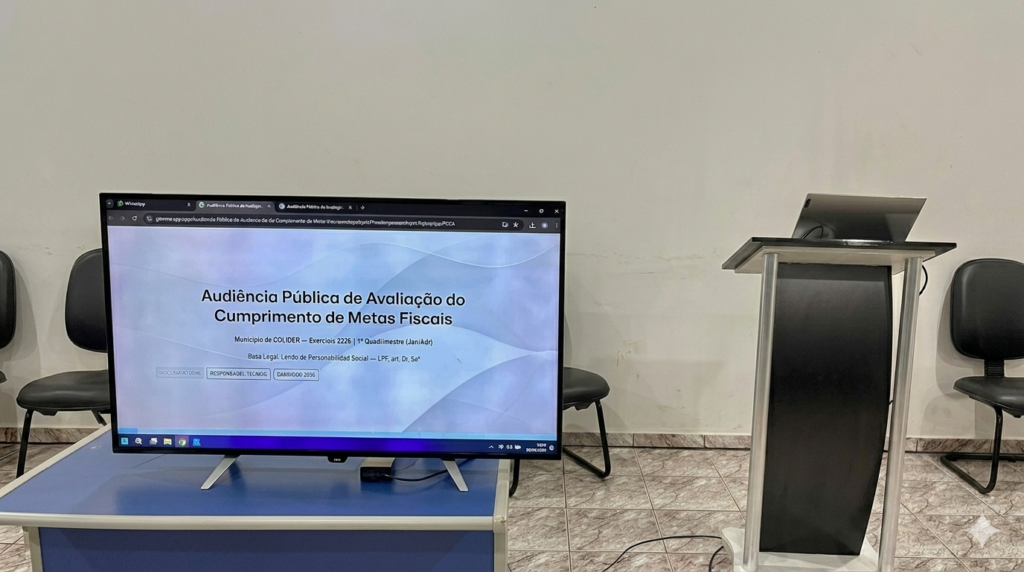 alt=Monitor de TV exibindo slide sobre Audiência Pública de Avaliação do Cumprimento de Metas Fiscais ao lado de um púlpito.